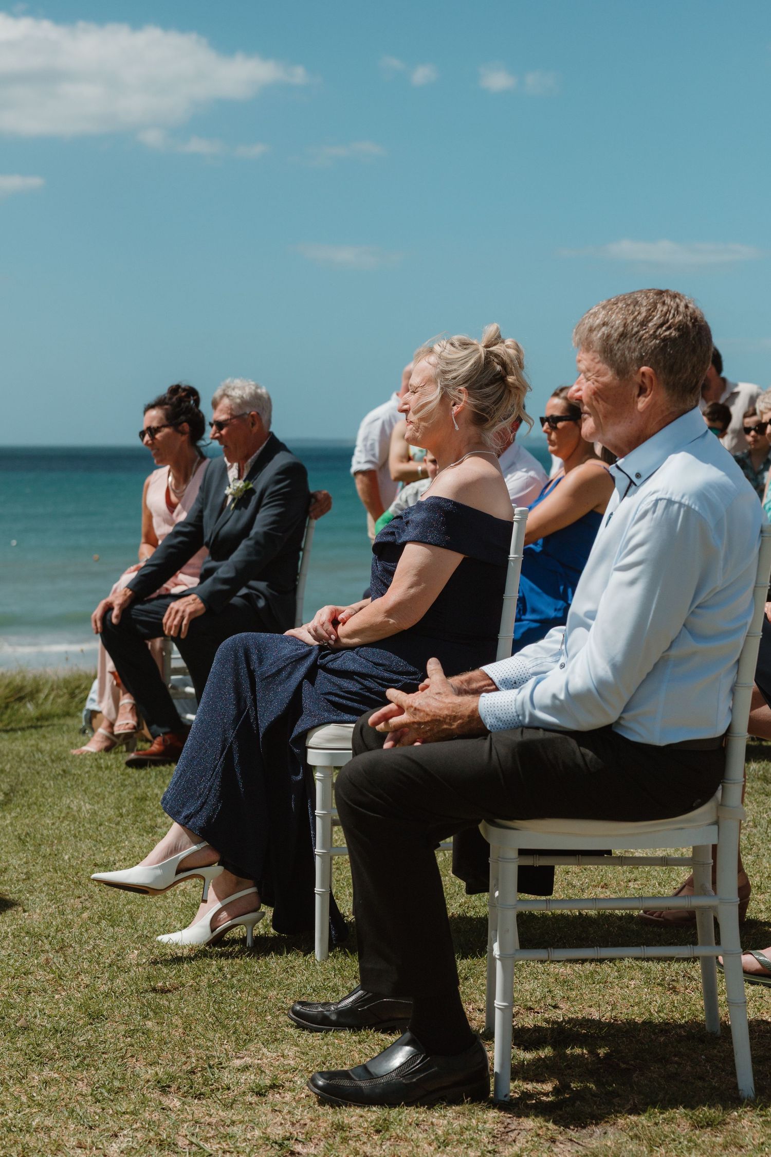 Guests seated on white chairs at an outdoor beach wedding ceremony on a sunny day.