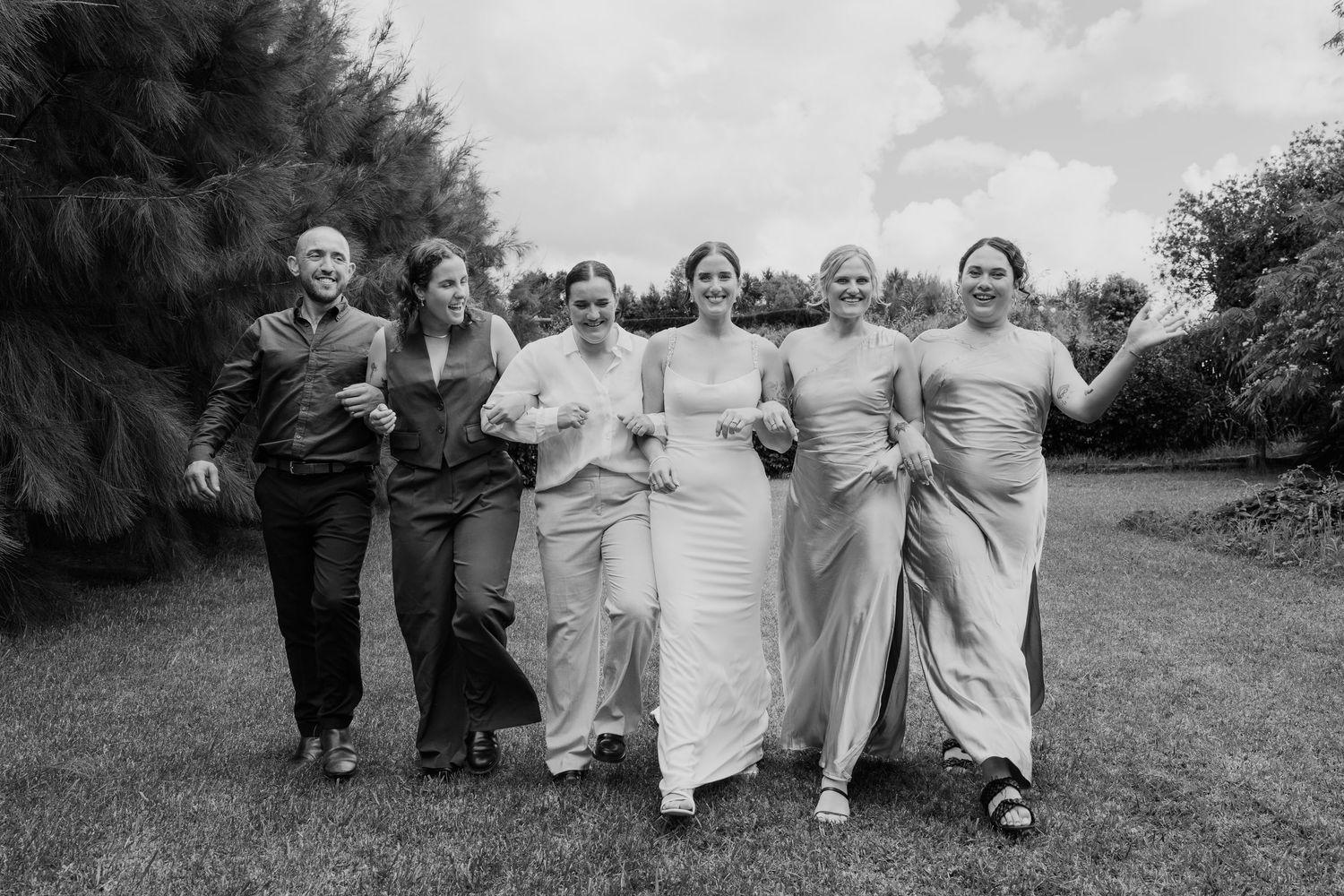 Black and white photo of a bride in white gown with five bridesmaids laughing and walking outdoors.