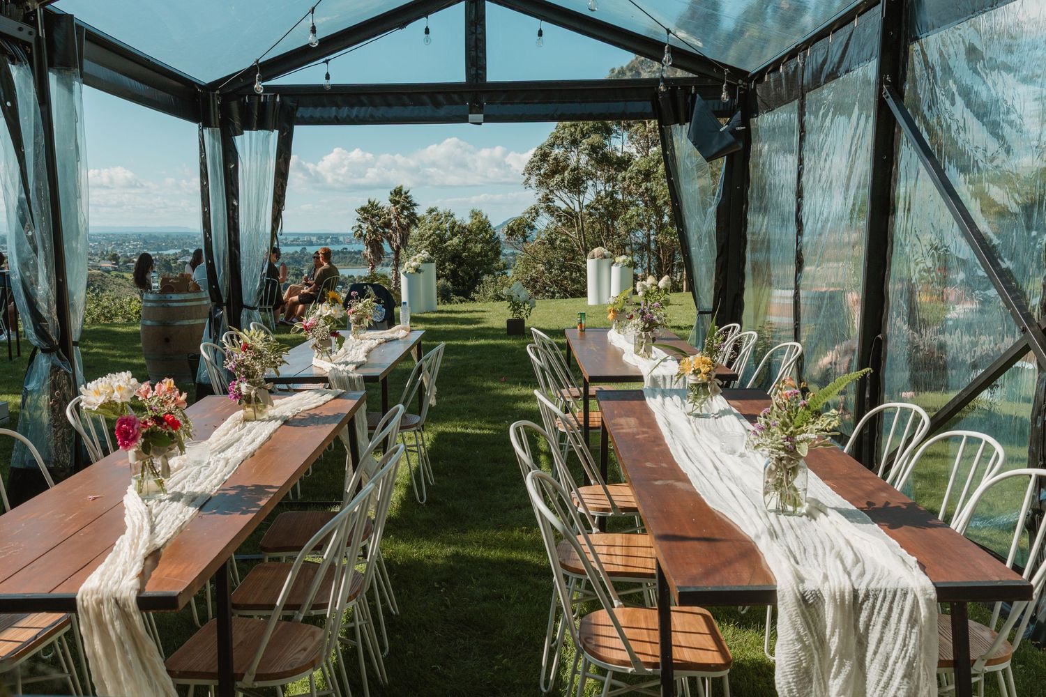 Elegant outdoor wedding reception setup with long wooden tables, white chairs, and floral centerpieces under a glass pavilion.