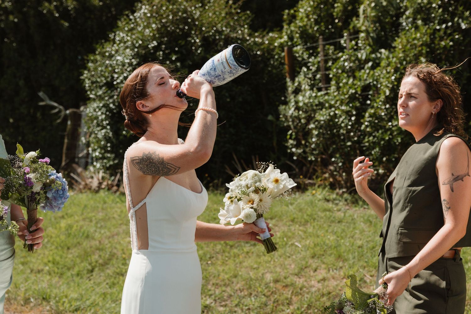 Bride in white dress drinking from water bottle while holding bouquet, with guest in green dress outdoors.