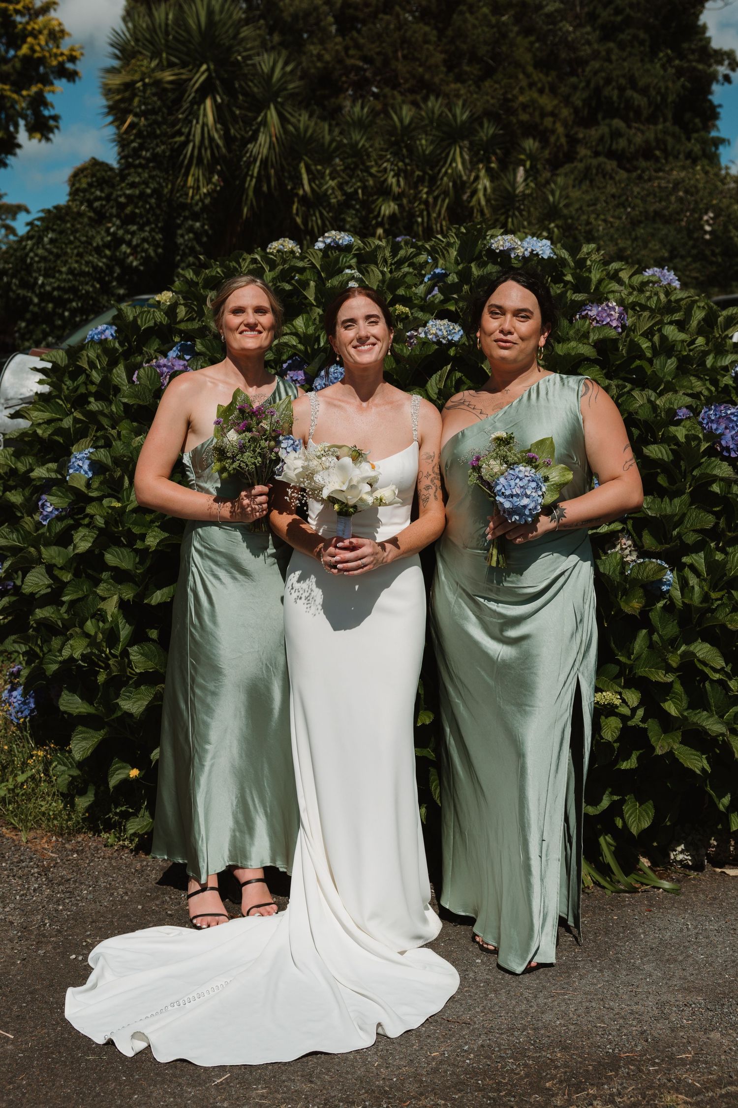 Bride in white gown with two bridesmaids in sage green satin dresses holding bouquets before hydrangea backdrop.