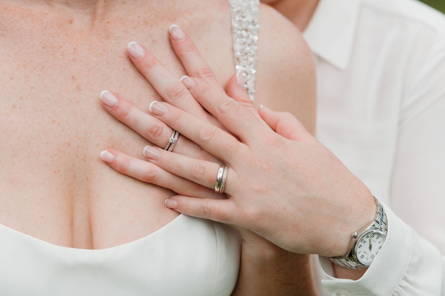 Close-up of newlyweds hands showing wedding bands, bride in white dress with groom wearing a silver watch.