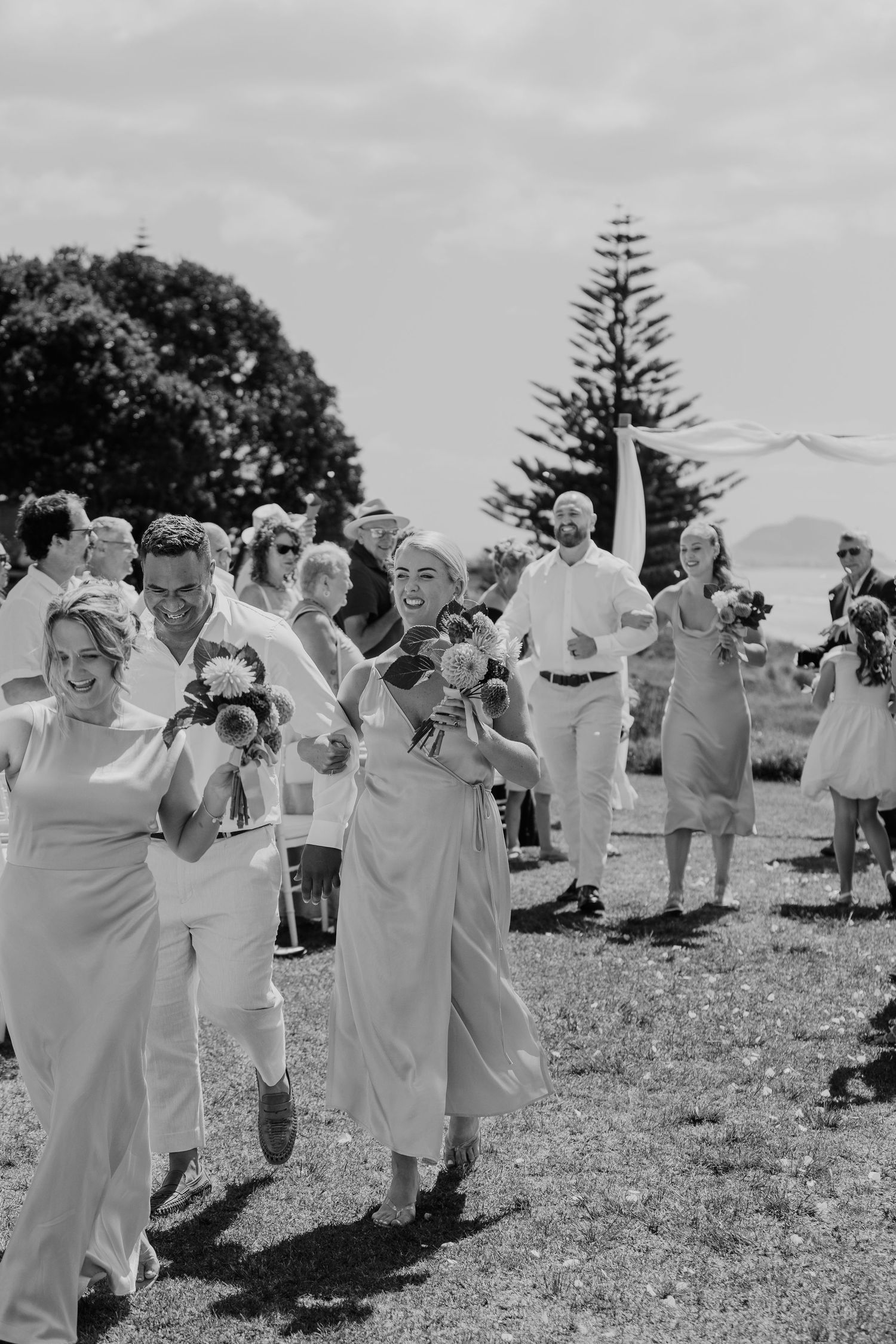 Black and white photo of joyful wedding guests walking outdoors, holding bouquets, smiling, with trees and sky in background.