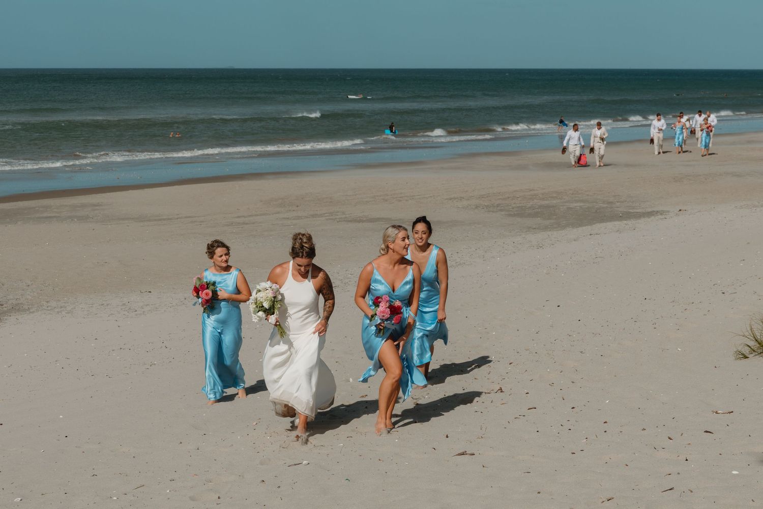 Bride in white gown walks with bridesmaids in blue dresses on a sunny beach during a wedding celebration.
