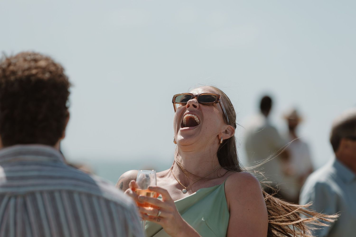 A joyful woman in sunglasses laughs outdoors at a beach gathering, holding a drink on a sunny day.