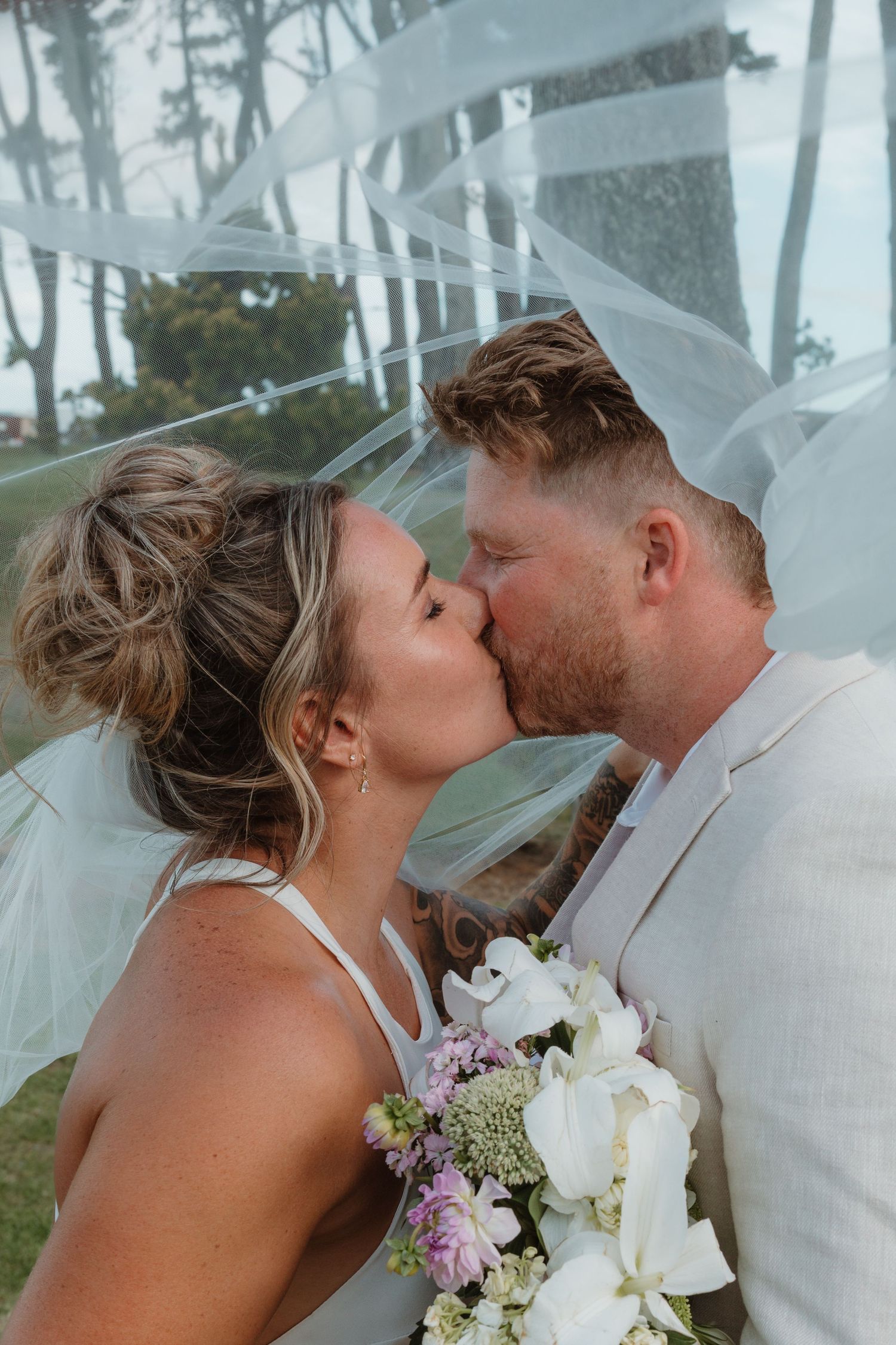 Bride and groom sharing a romantic kiss under a flowing white veil, bride holding a white and purple floral bouquet.