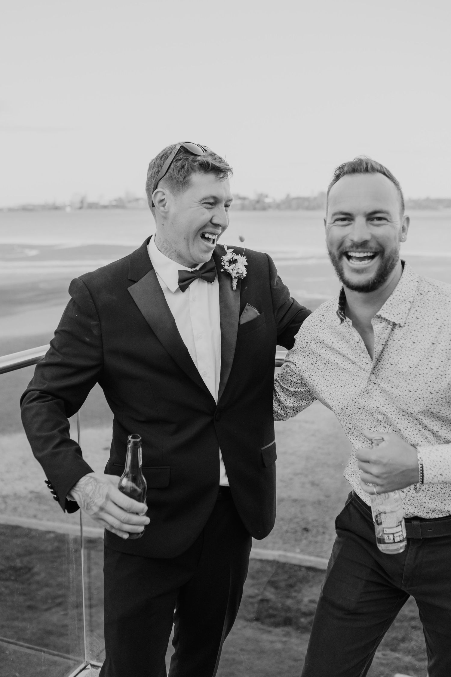 Two friends share a joyful moment outdoors, laughing together while holding drinks at a formal event by the water.