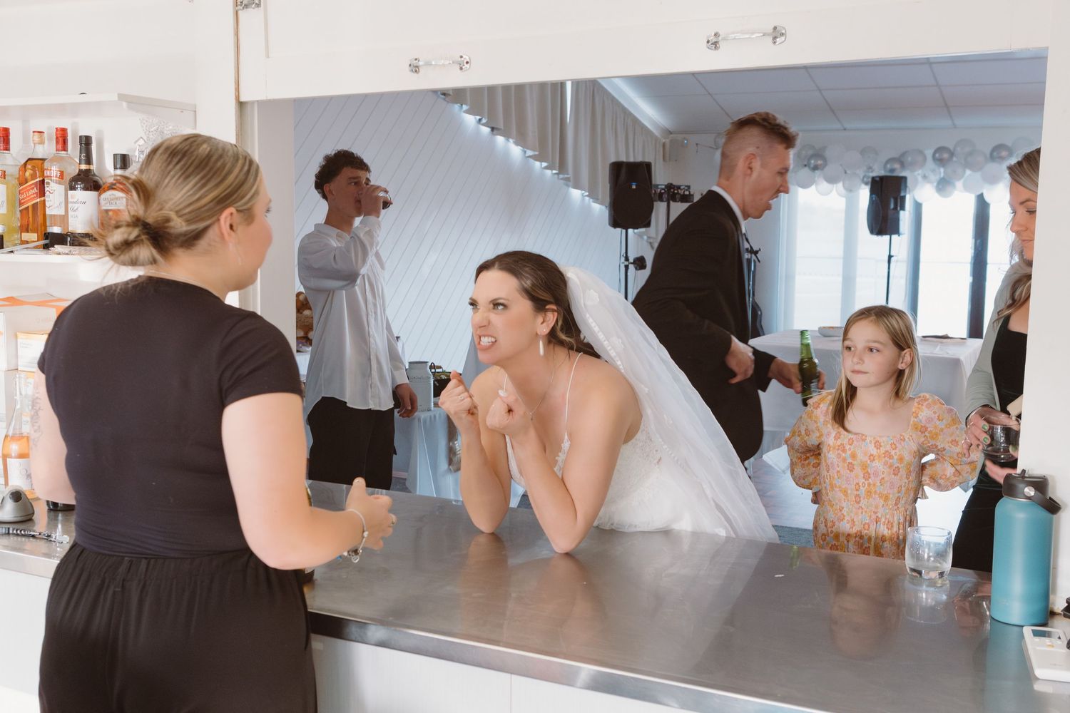 Inside view of a wedding reception bar area shows people interacting in a bright white venue space.