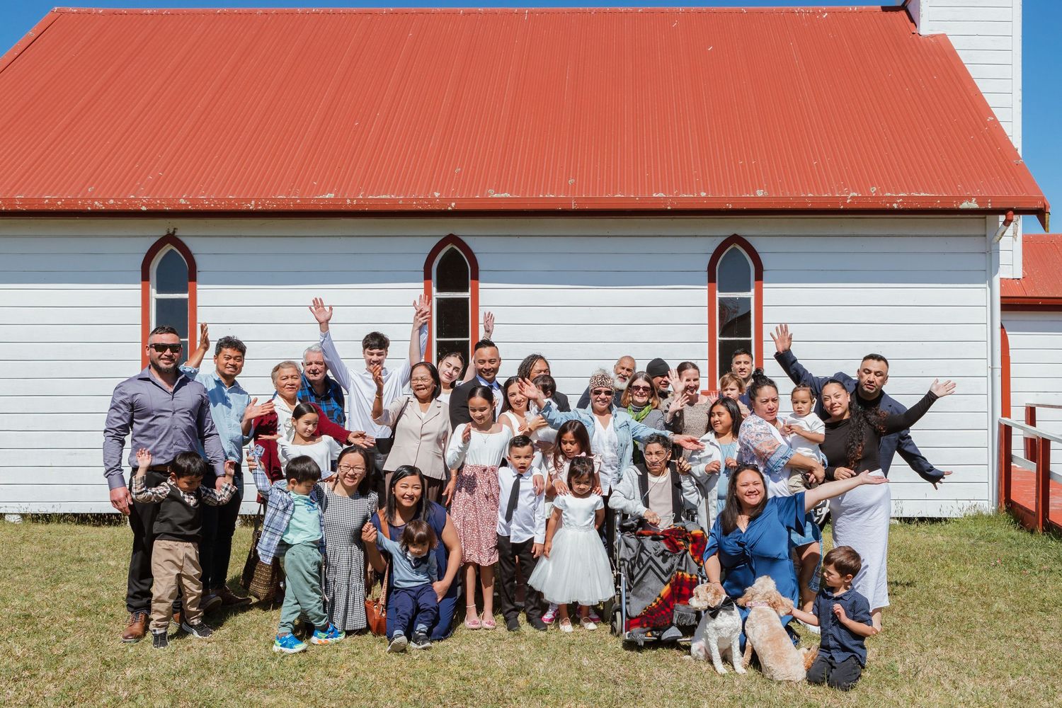 Large group poses joyfully outside white church building with red roof and gothic style windows on sunny day.