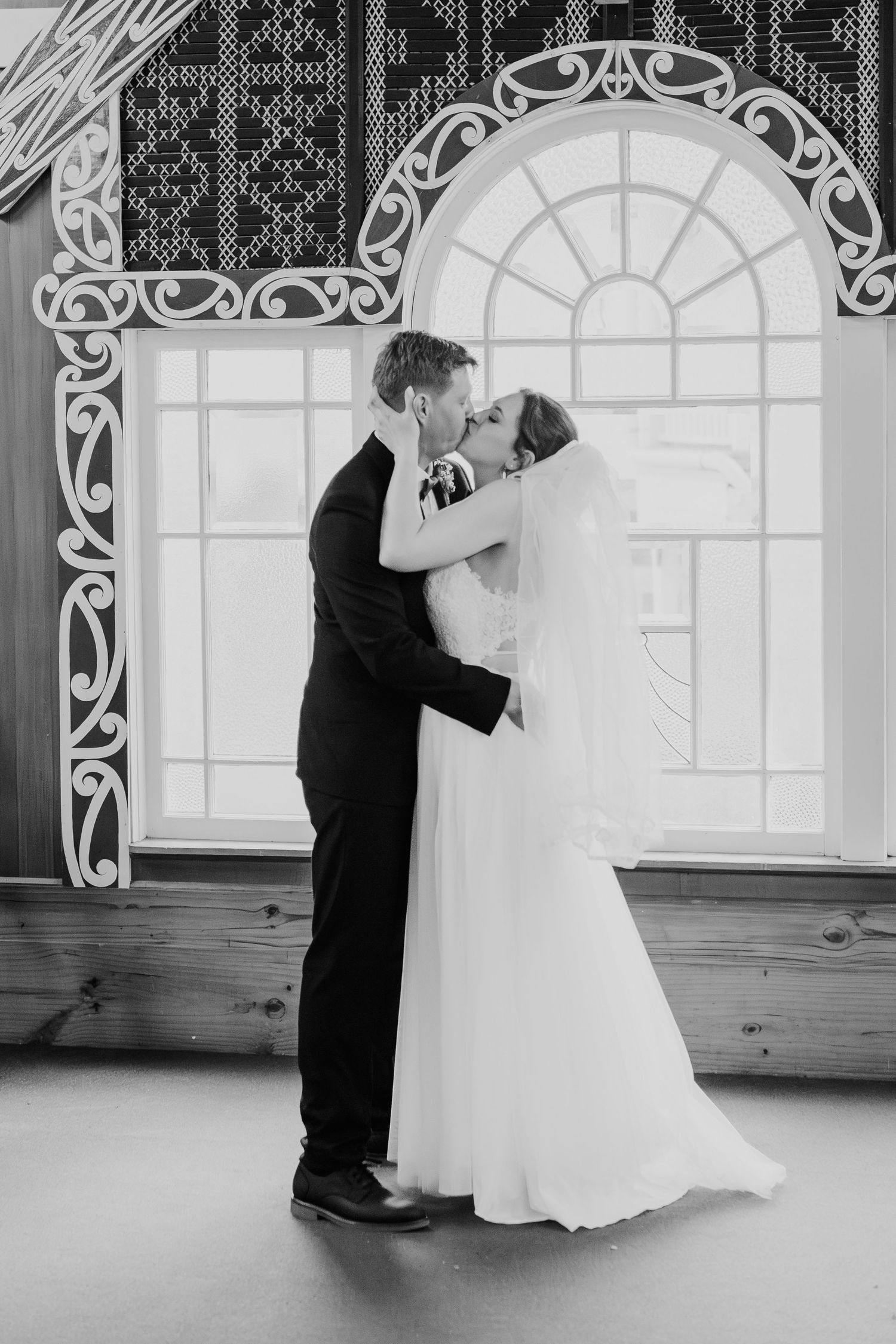 A romantic black and white wedding photograph of a couple sharing their first kiss in front of an ornate arched window.