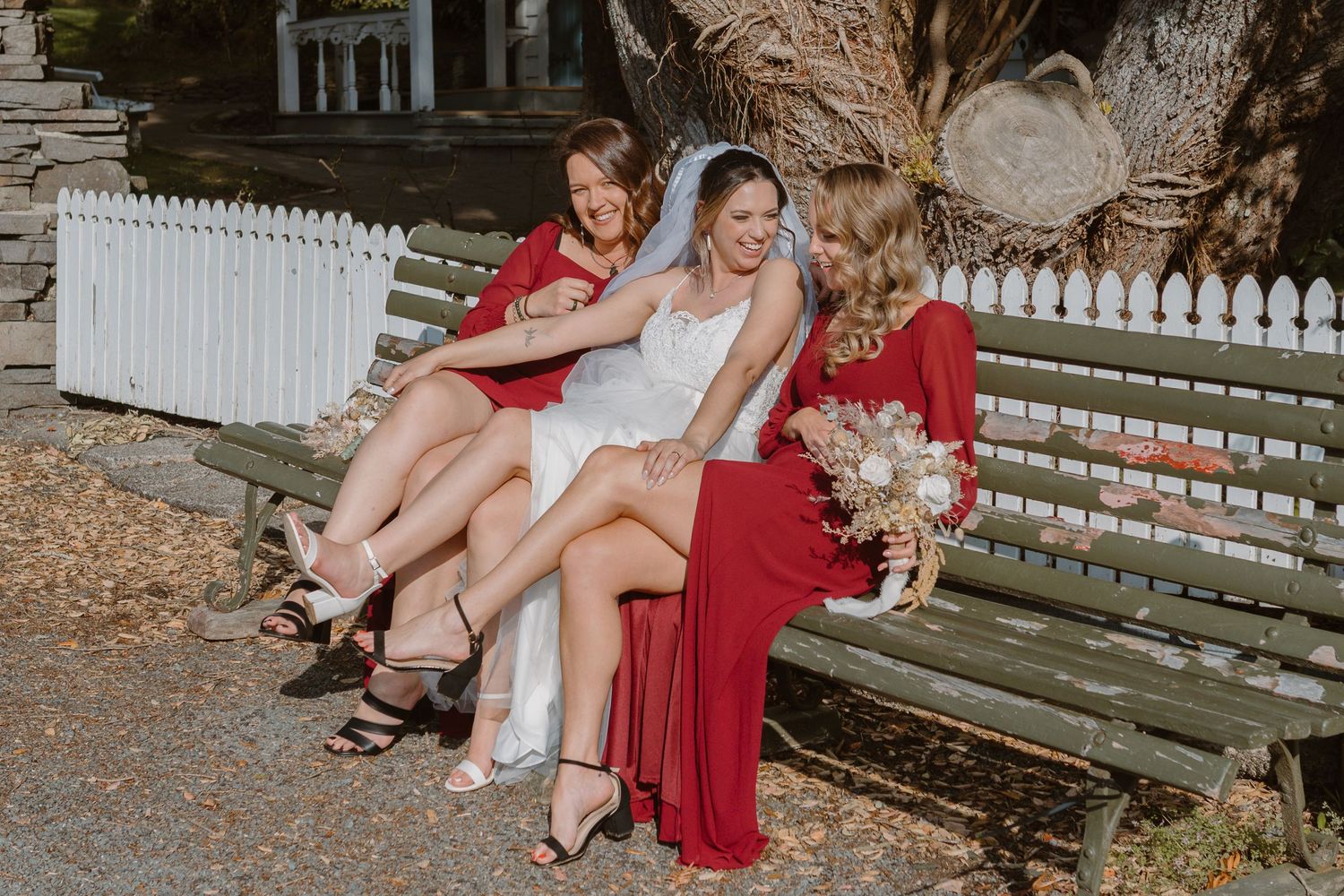 Three people in formal red and white attire sitting casually on a green park bench by a white picket fence.