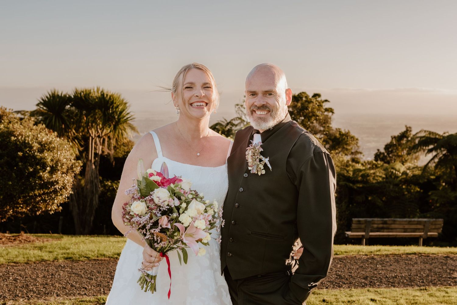 Two people stand together outdoors at sunset, one in a white wedding dress holding a bouquet and one in dark formal attire.