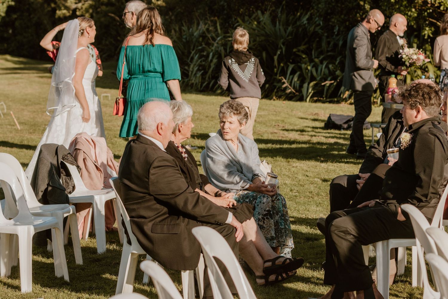 Wedding guests sit in white chairs on a sunny lawn during an outdoor ceremony at sunset.
