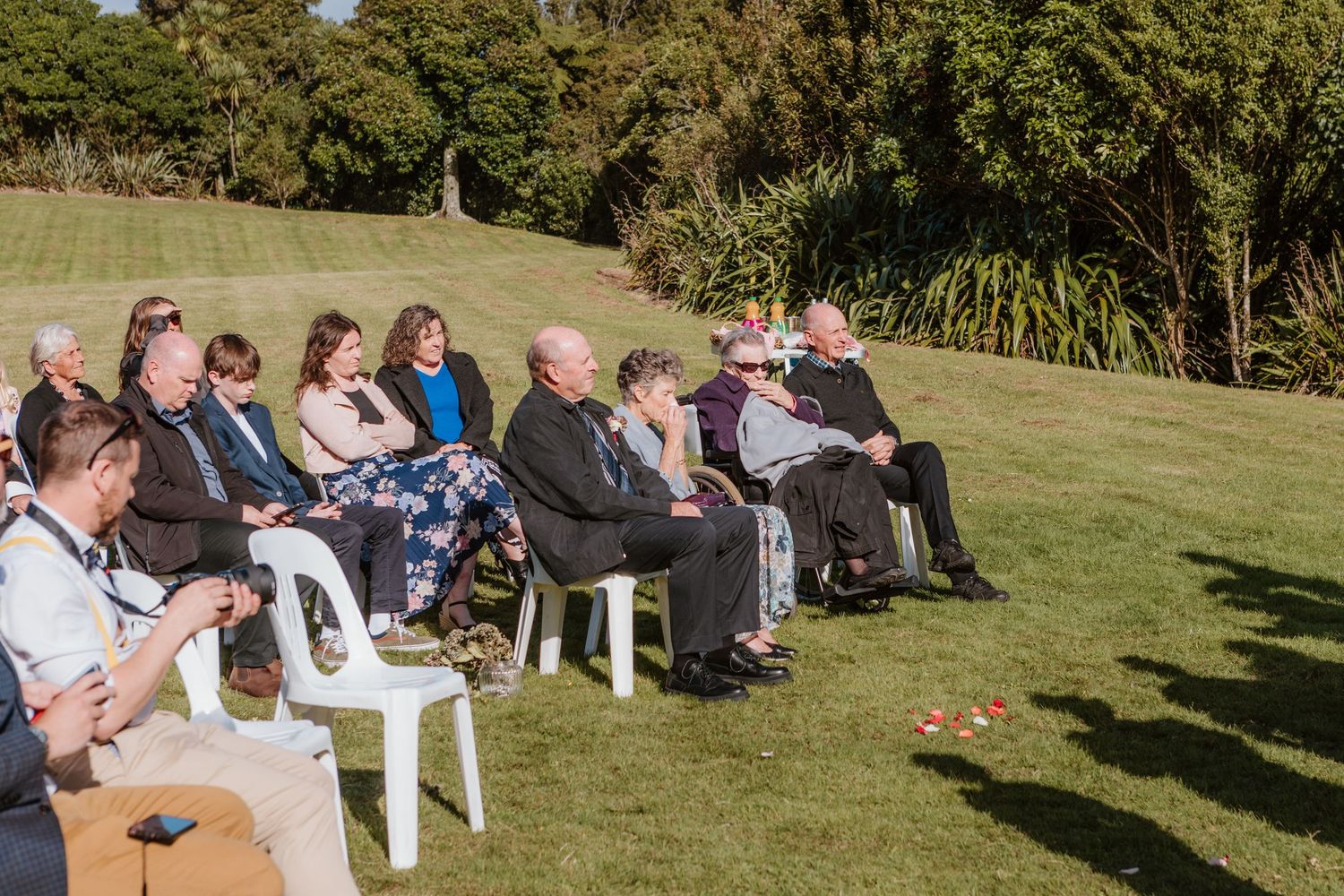 Wedding guests sit in white chairs on a grassy lawn during an outdoor ceremony in bright sunlight.