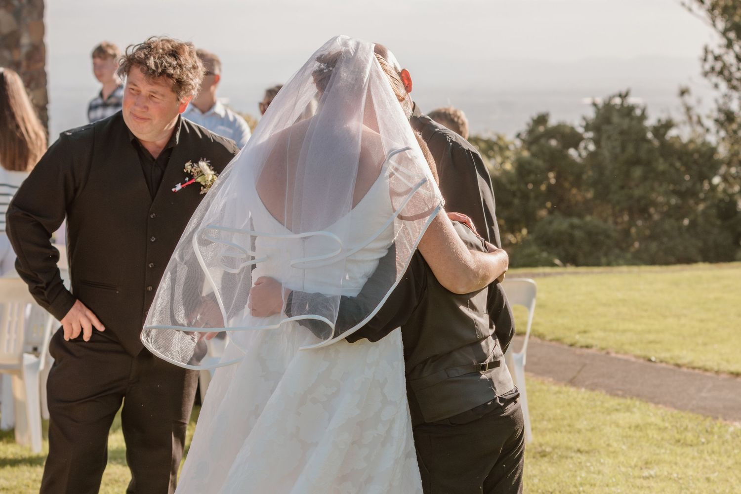 Wedding party gathers outdoors in sunlight on grassy hillside with a bride wearing a flowing white veil and dress.