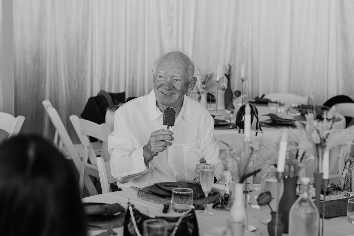 A gentleman in white clothing smiles while smoking at a table with wine bottles and table settings in black and white.