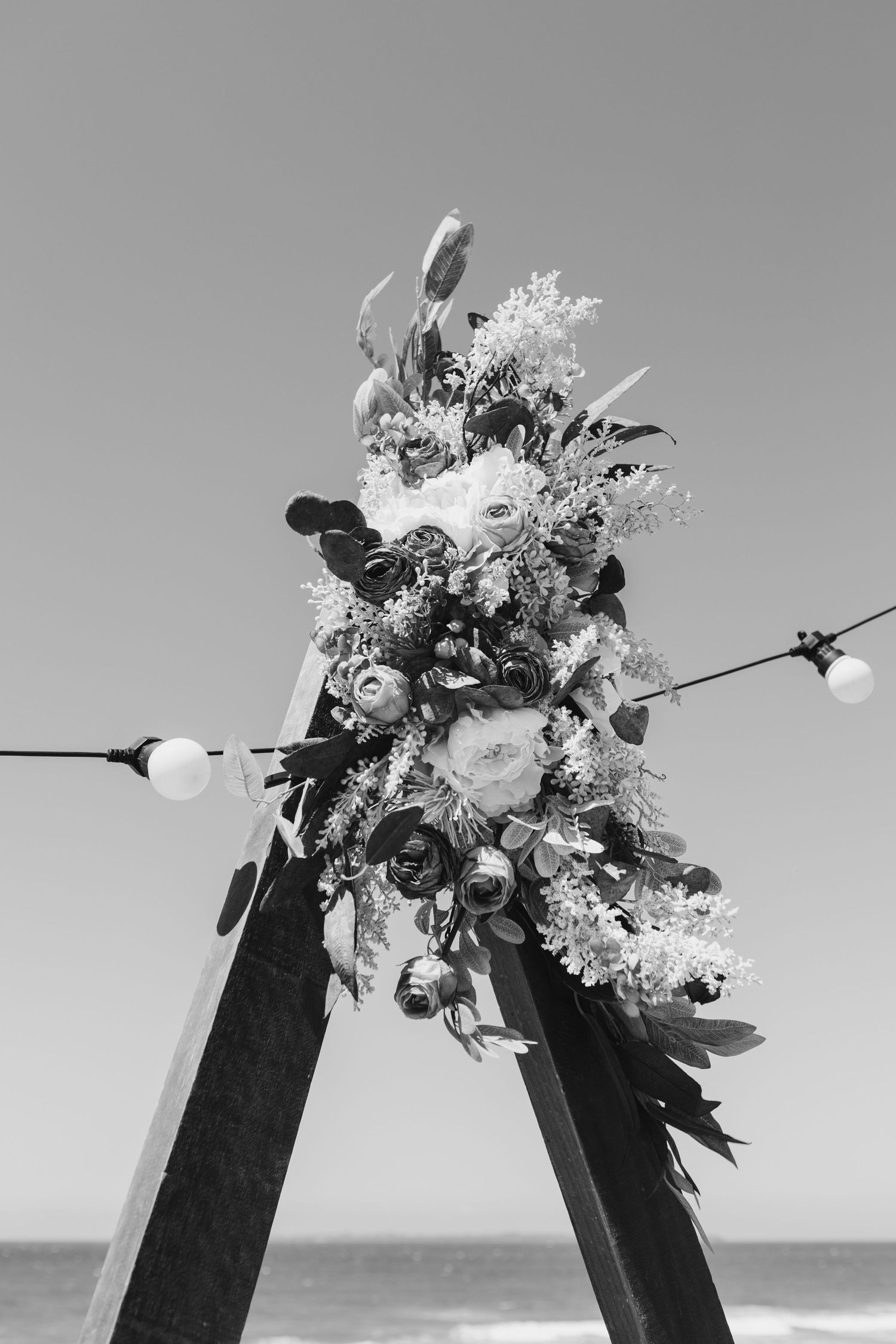 A wooden wedding arch decorated with white and pink flowers stands against a beach backdrop in black and white.