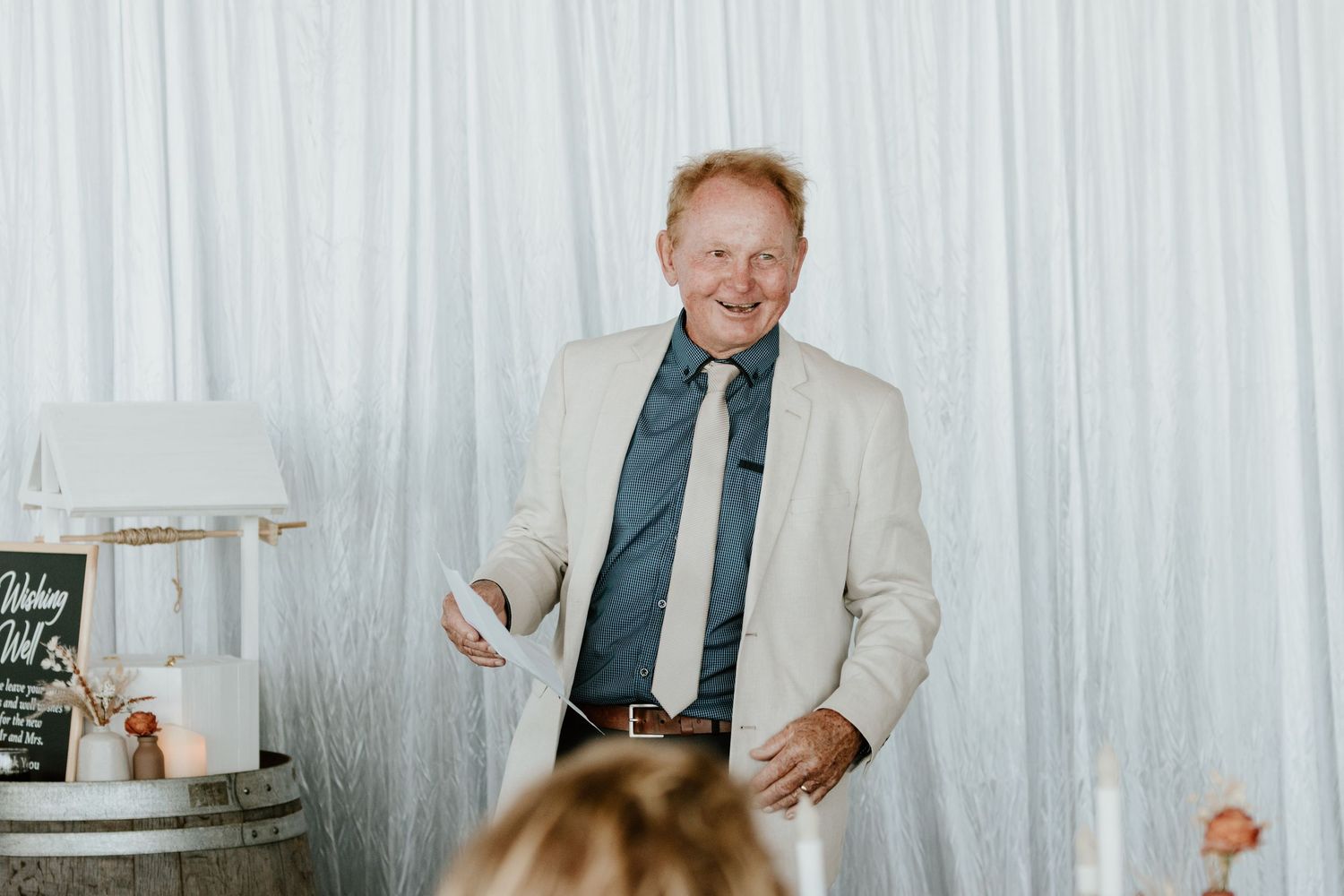 A person in a light suit and blue tie smiles while giving a speech at an event with white drapes in the background.