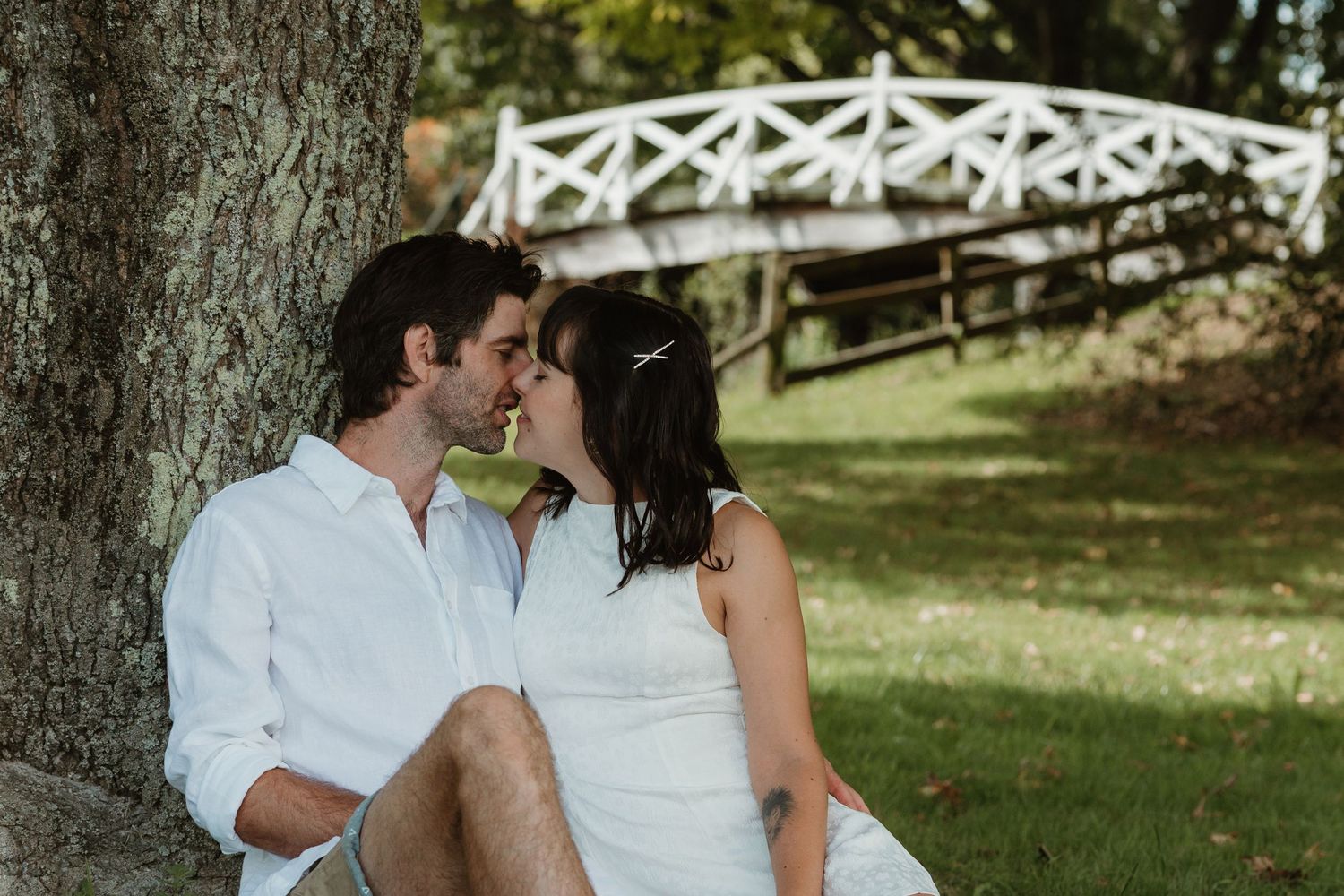 Couple sharing a romantic almost-kiss while sitting against a tree trunk in a lush green park with a white bridge backdrop.