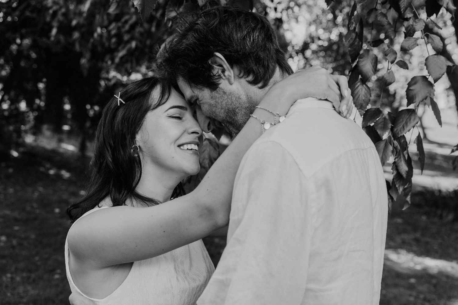 Black and white photo of a smiling couple sharing a romantic moment under leafy branches outdoors.