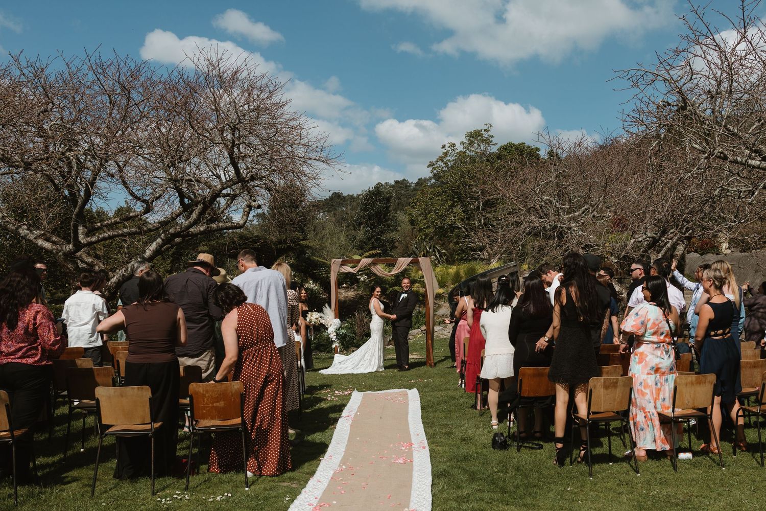 Outdoor wedding ceremony with wooden chairs and guests gathered under bare trees on a sunny day.