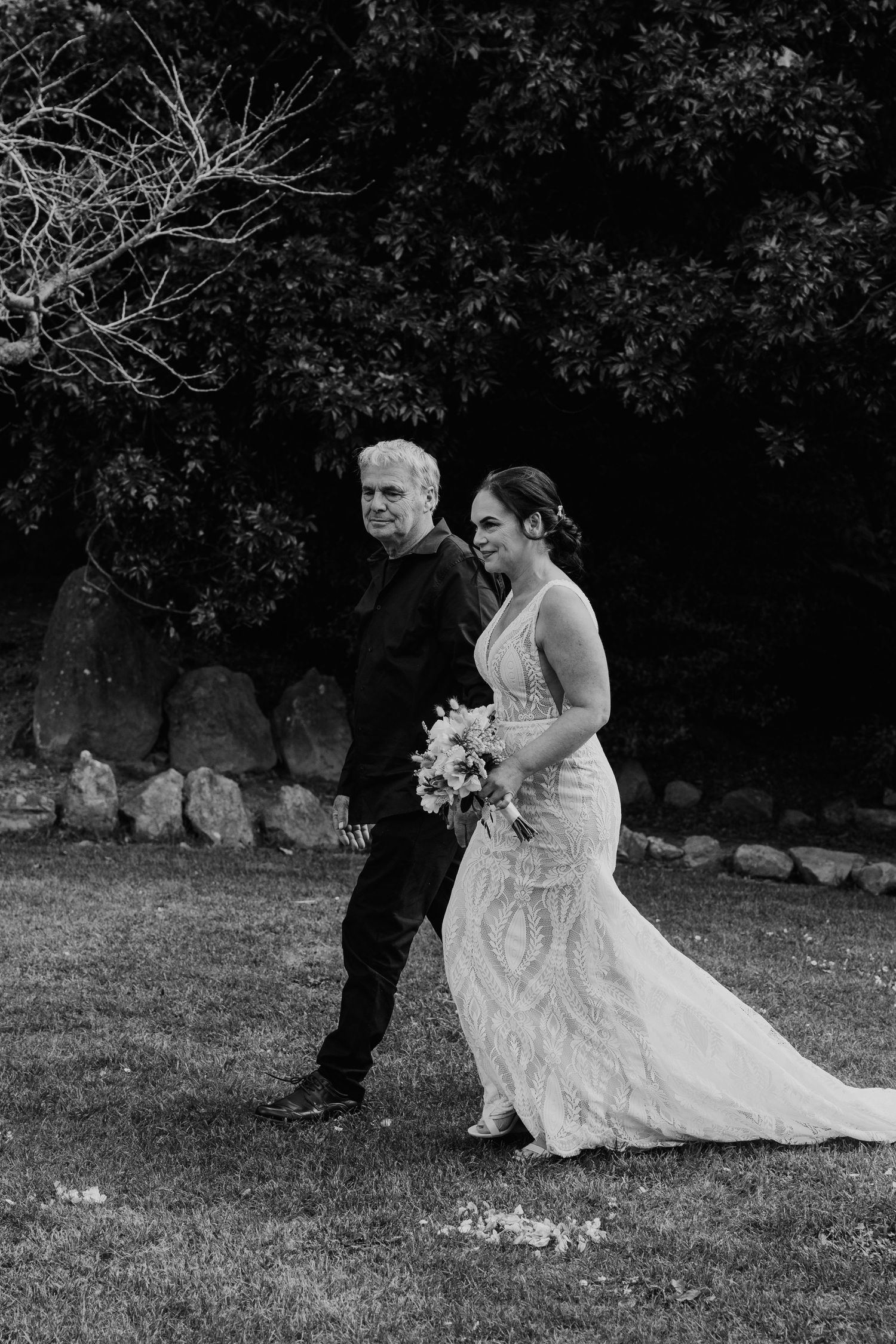 A black and white wedding photo showing a bride in a lace gown walking with a companion on a grassy lawn.