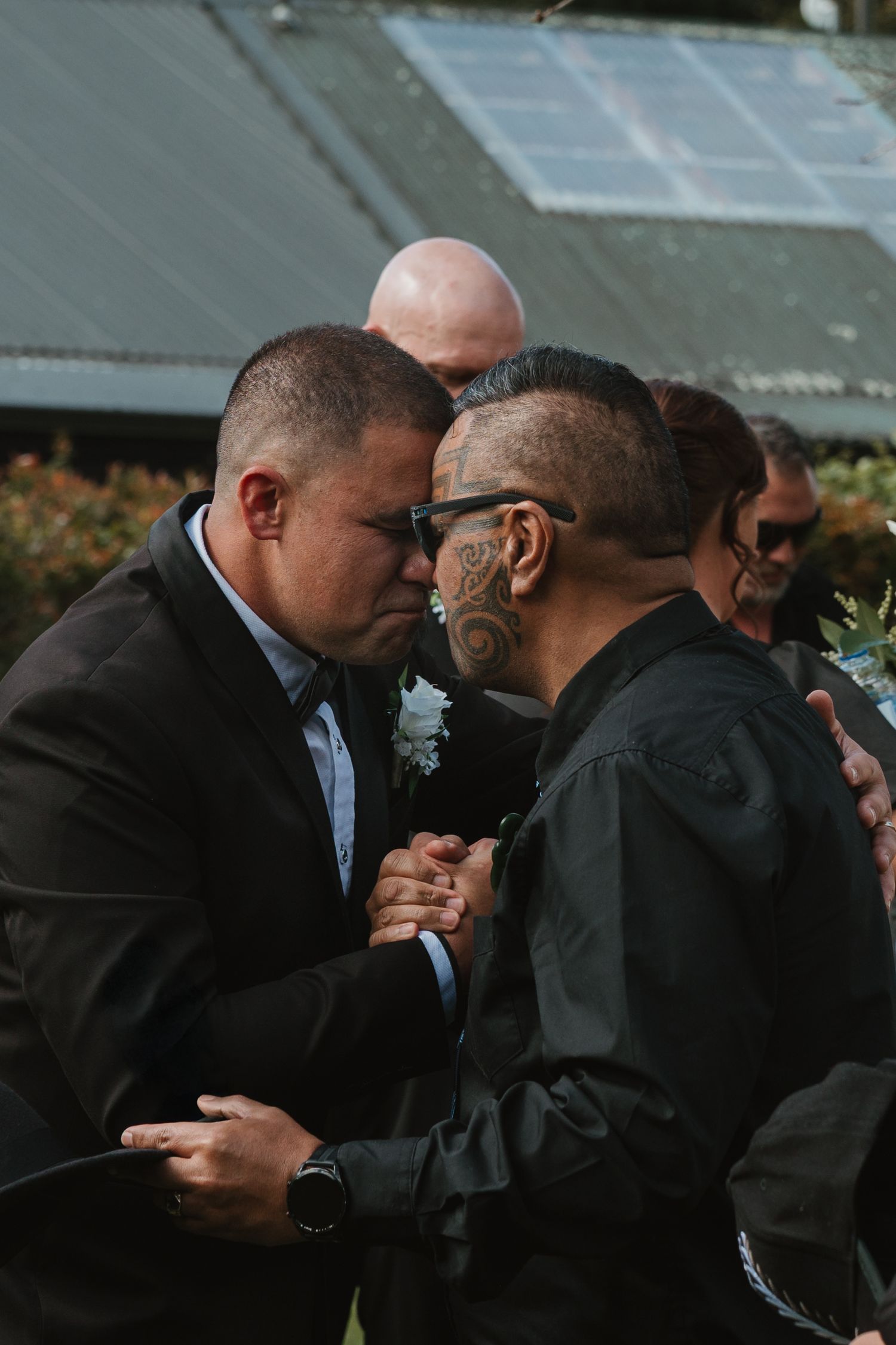 Two men in dark formal attire embrace and share an emotional moment during an outdoor event.