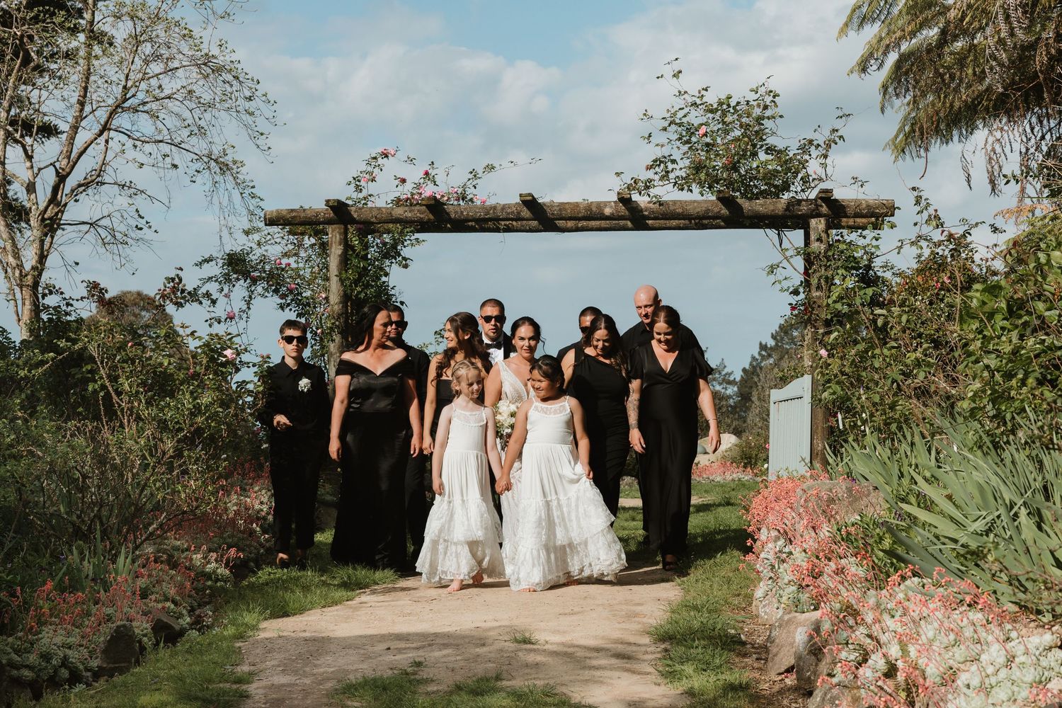 Wedding party walking along a garden path lined with trees and flowering plants on a sunny day.