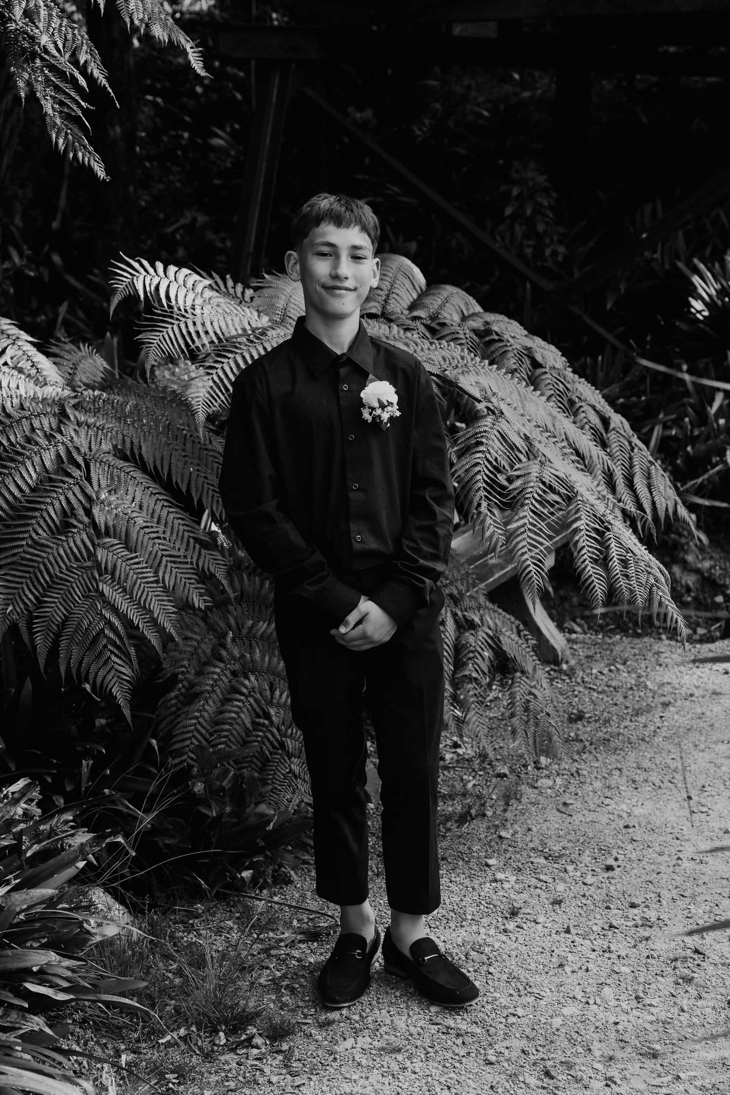 A figure in formal black attire stands along a garden path surrounded by lush ferns and greenery.