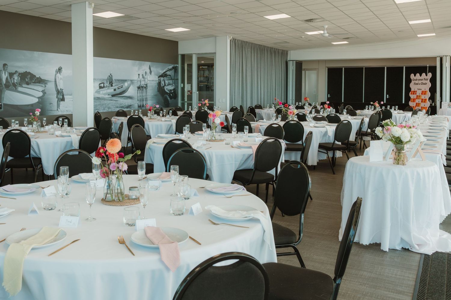 Elegant banquet hall with round tables draped in white linens, black chairs, and colorful floral centerpieces for a wedding reception.