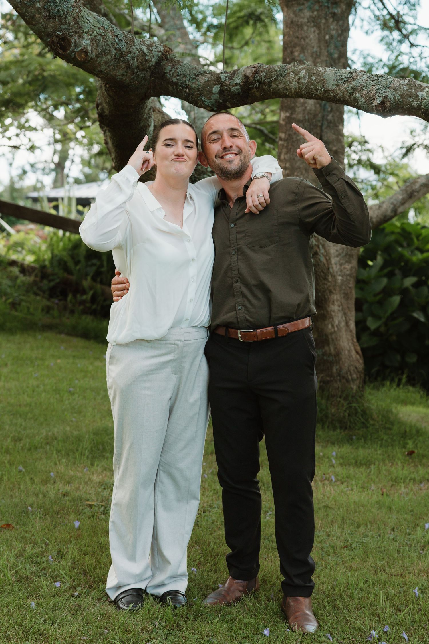 Two smiling friends pose together outdoors under a large tree, one in white outfit and one in olive shirt.