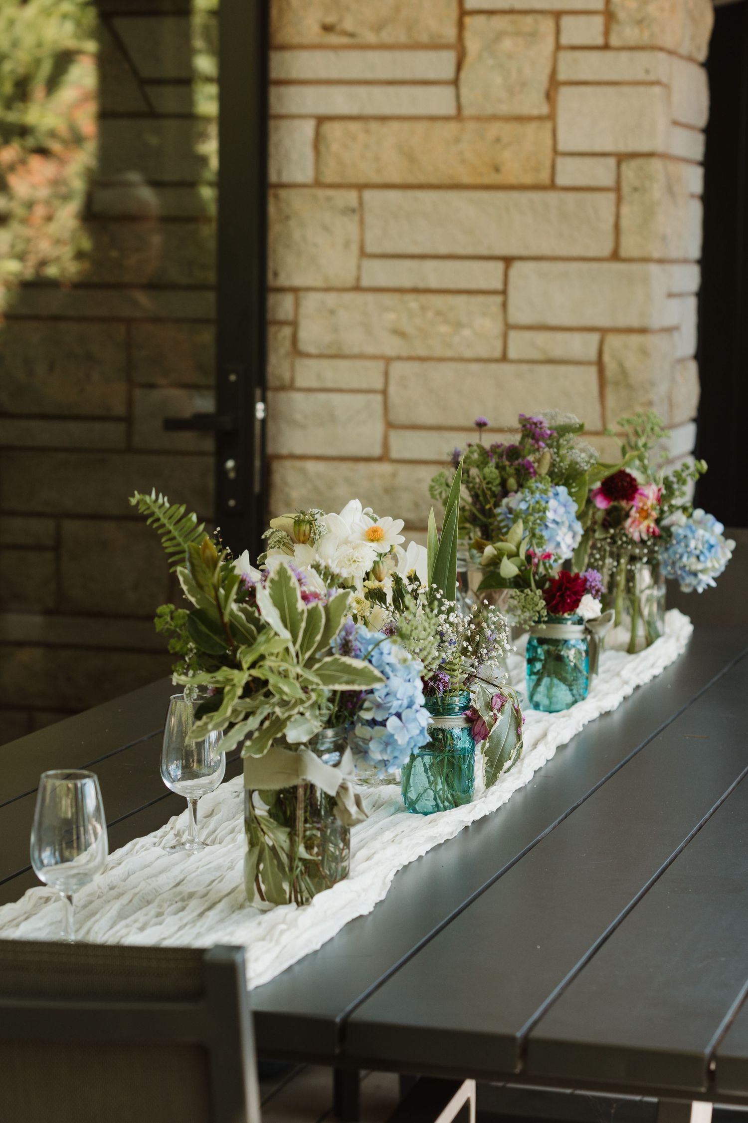 Rustic outdoor table with mason jar floral centerpieces featuring blue hydrangeas and wildflowers on a marble runner.