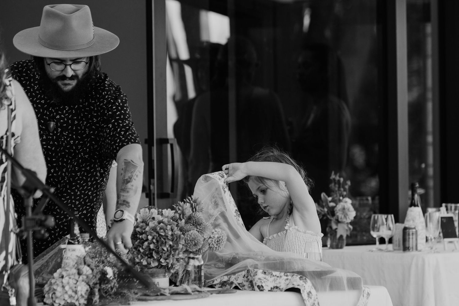 Black and white photo of tattooed man in wide-brim hat with young girl arranging flowers at wedding table.