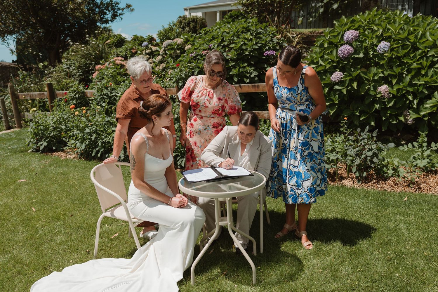 Bride in white gown signs documents at garden table with three women witnesses on sunny wedding day.