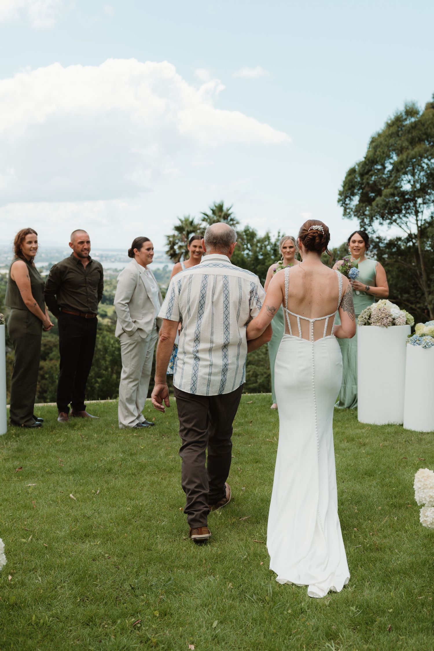 Bride in white gown walks with her father down the aisle at an outdoor wedding ceremony on a lush green lawn.