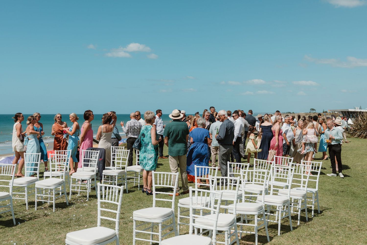 Outdoor beach wedding ceremony with white chairs and guests gathered on green lawn overlooking the ocean.