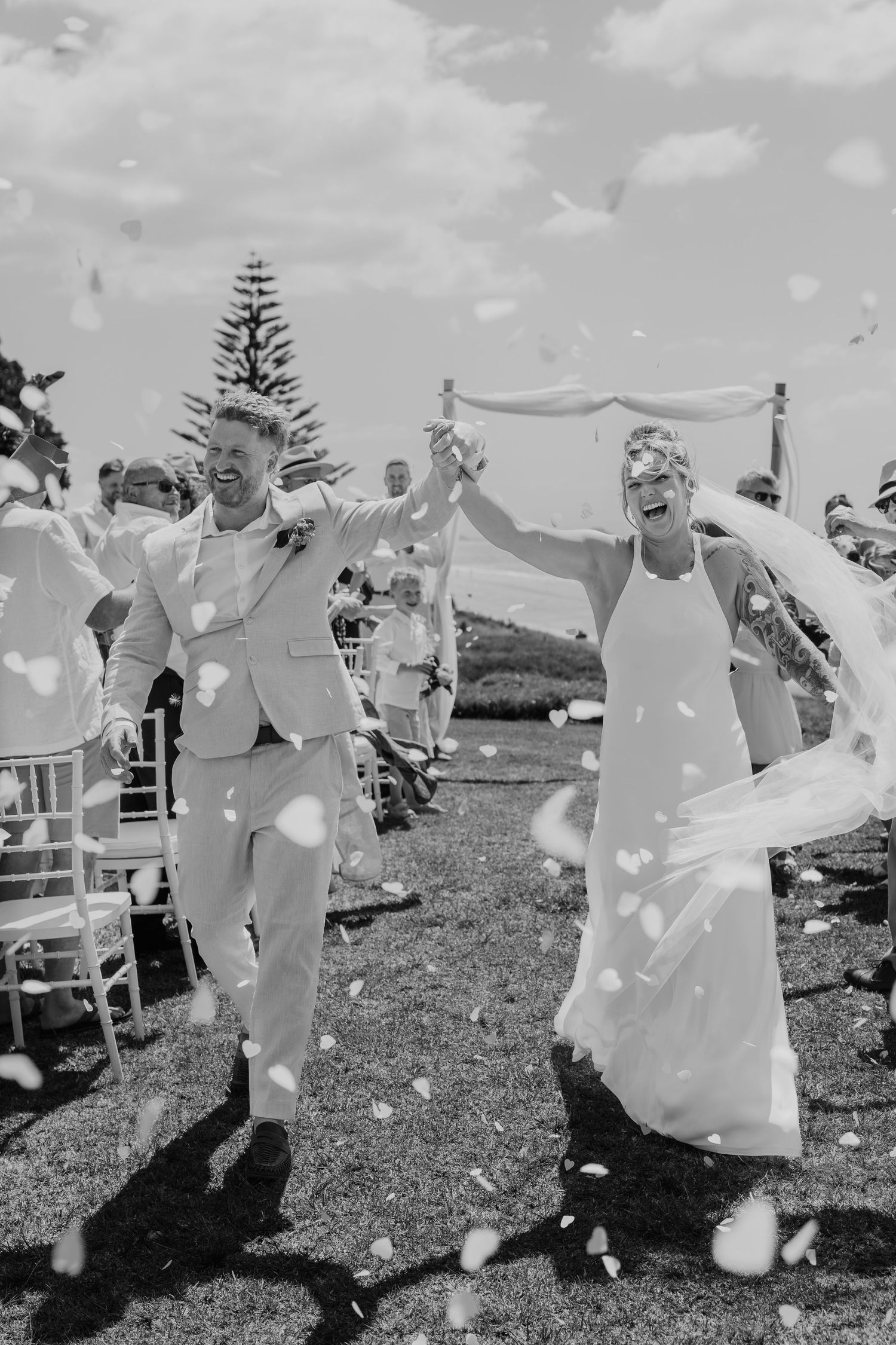Black and white photo of joyful newlyweds walking down the aisle as confetti fills the air at an outdoor wedding.