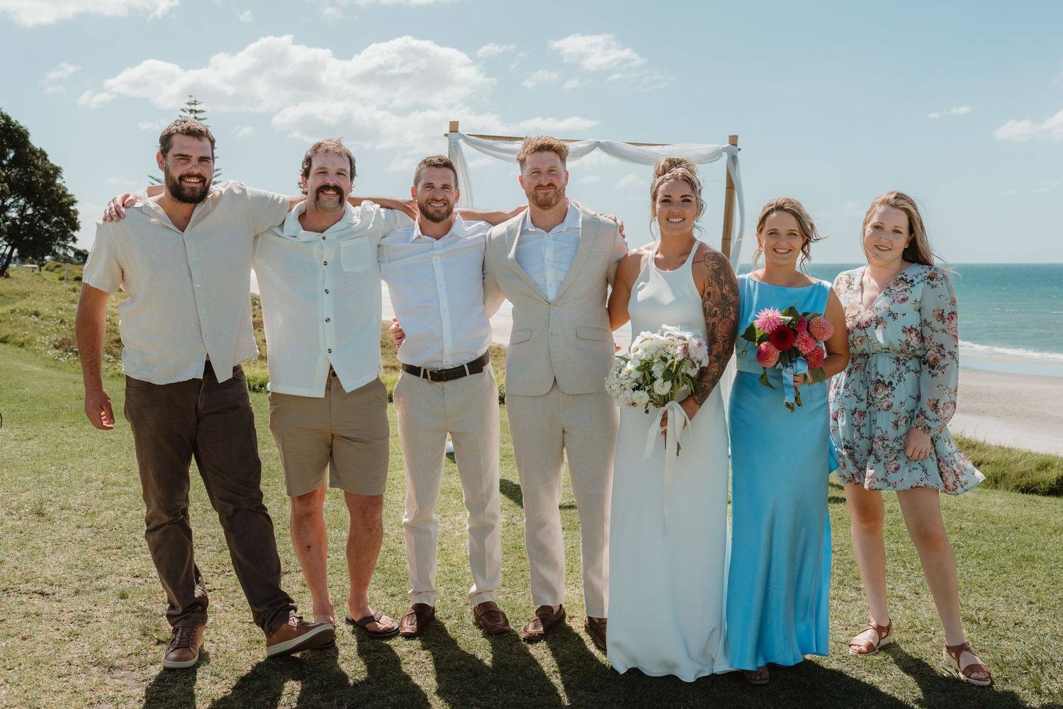 Wedding party posing outdoors by the ocean, featuring bride in white, groom in tan suit, and guests in formal attire.