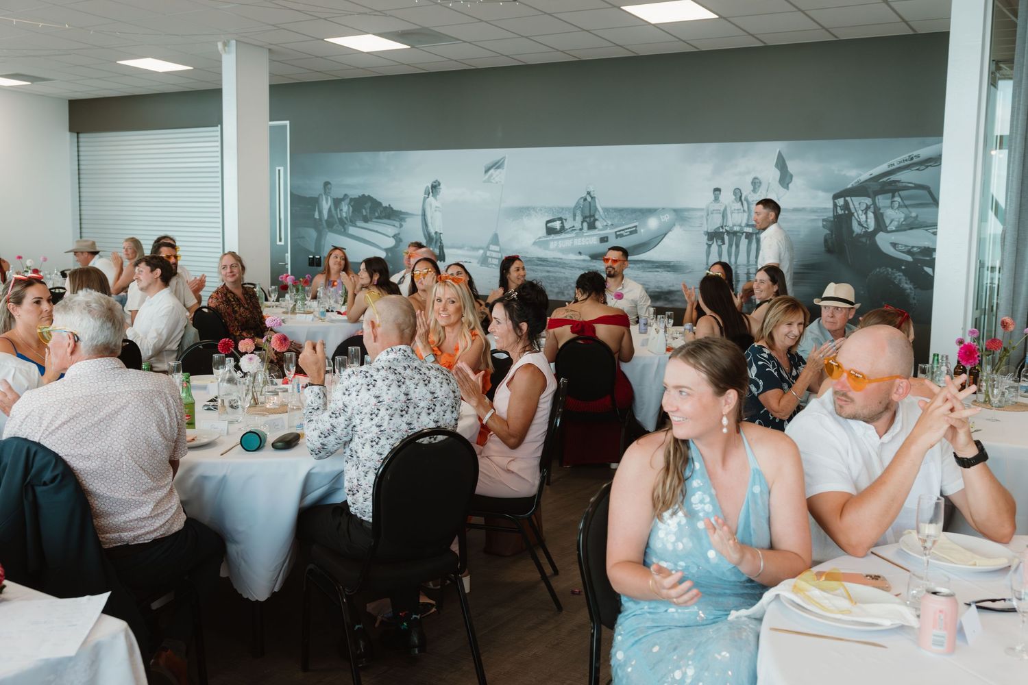 Guests enjoying a wedding reception dinner in a modern venue with white tablecloths and a scenic mural backdrop.