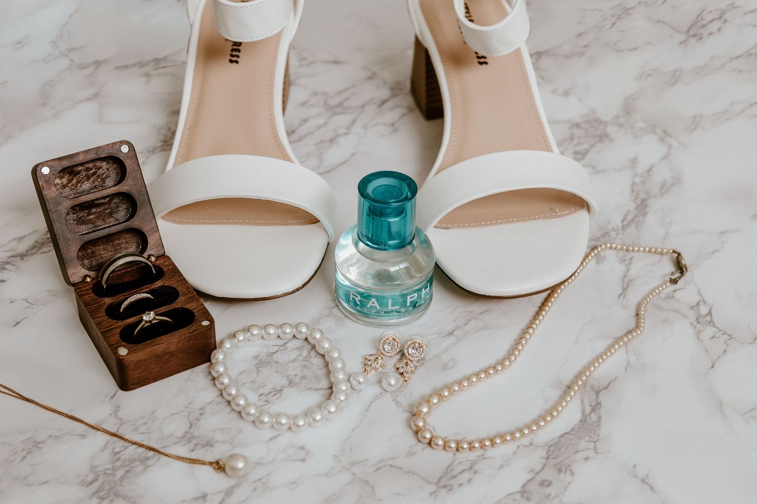 Wedding accessories including white heels, perfume, jewelry and rings displayed on marble surface.
