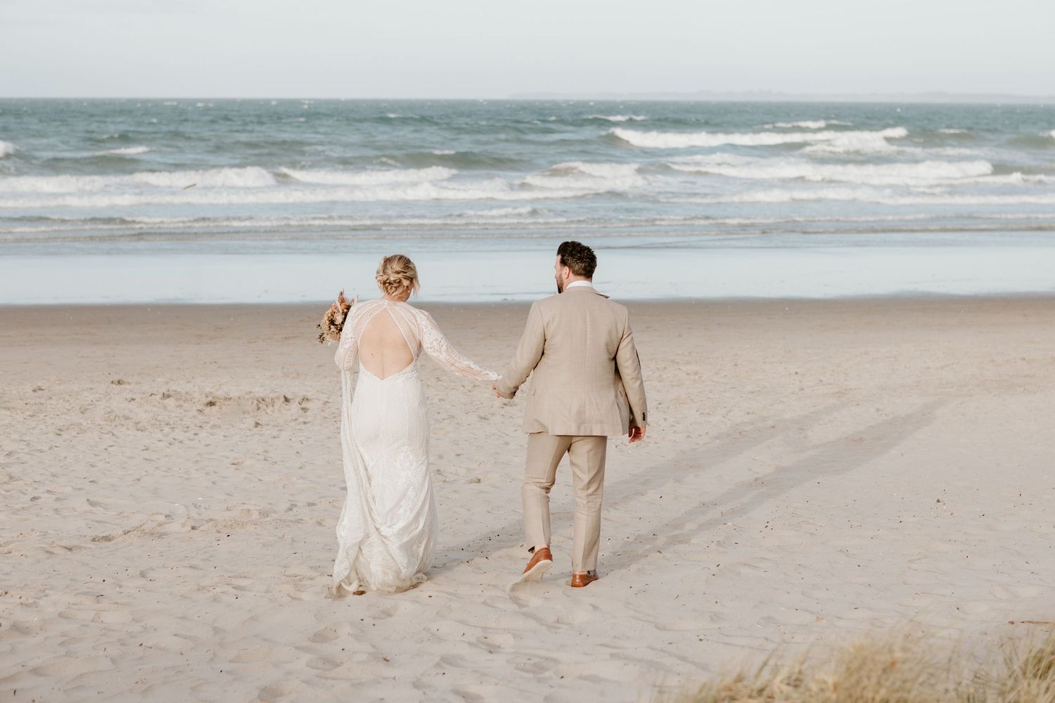 A couple in wedding attire walks hand in hand along a windy beach with ocean waves crashing in the background.