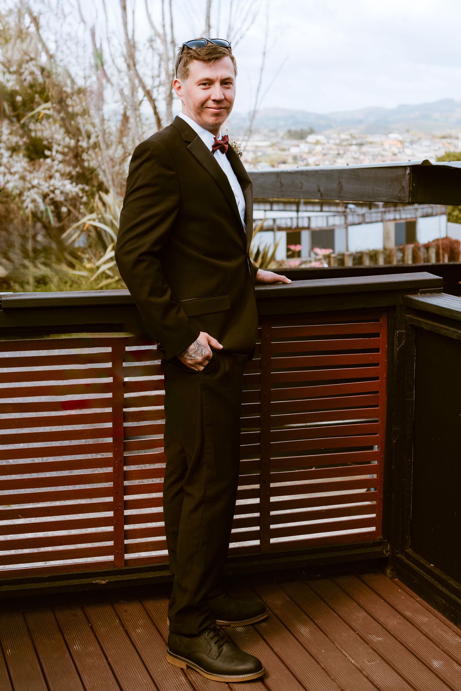 Someone wearing a classic black tuxedo poses on a wooden deck with mountain views in the background.