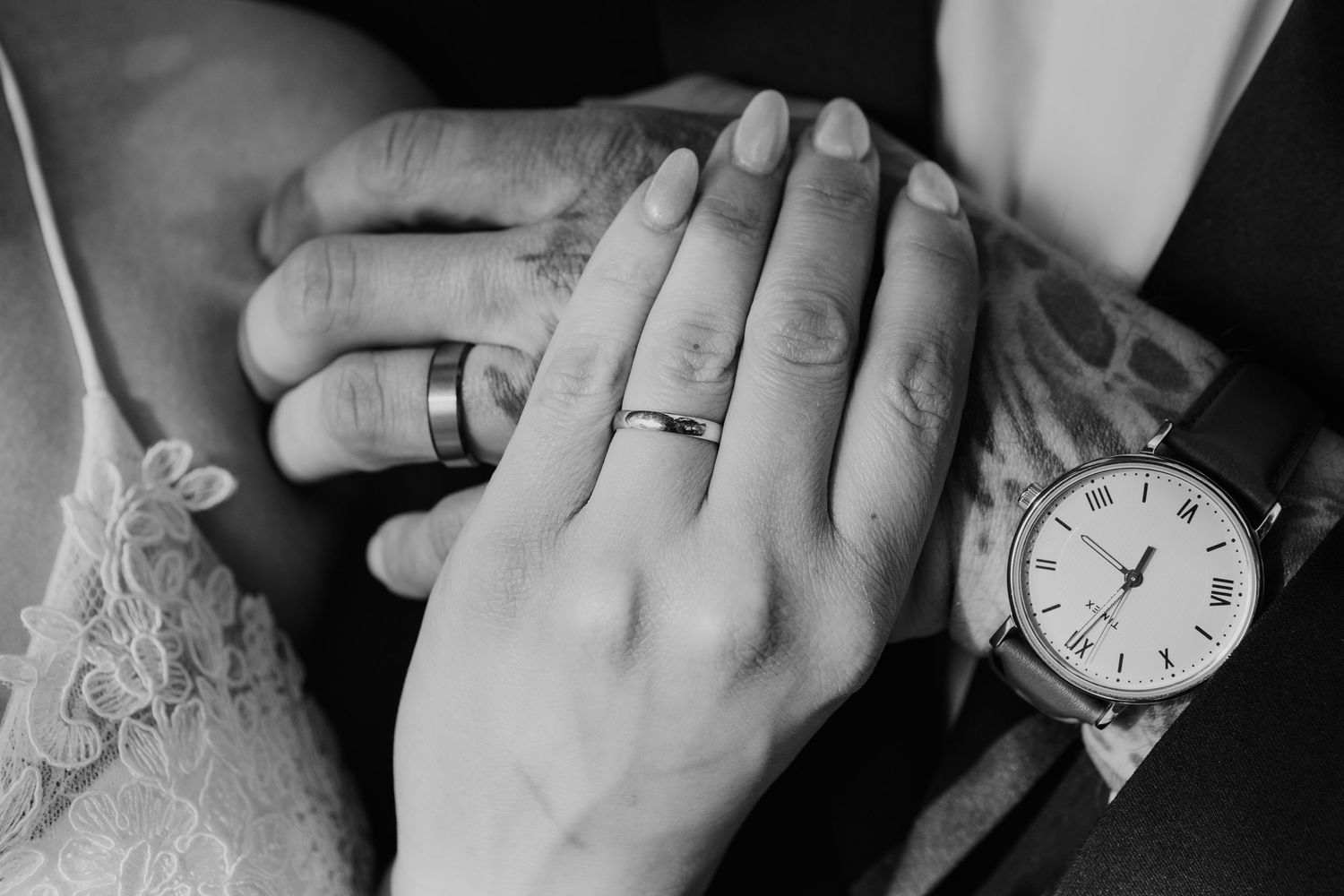 A black and white close-up of hands with wedding rings and a wristwatch against white lace fabric.