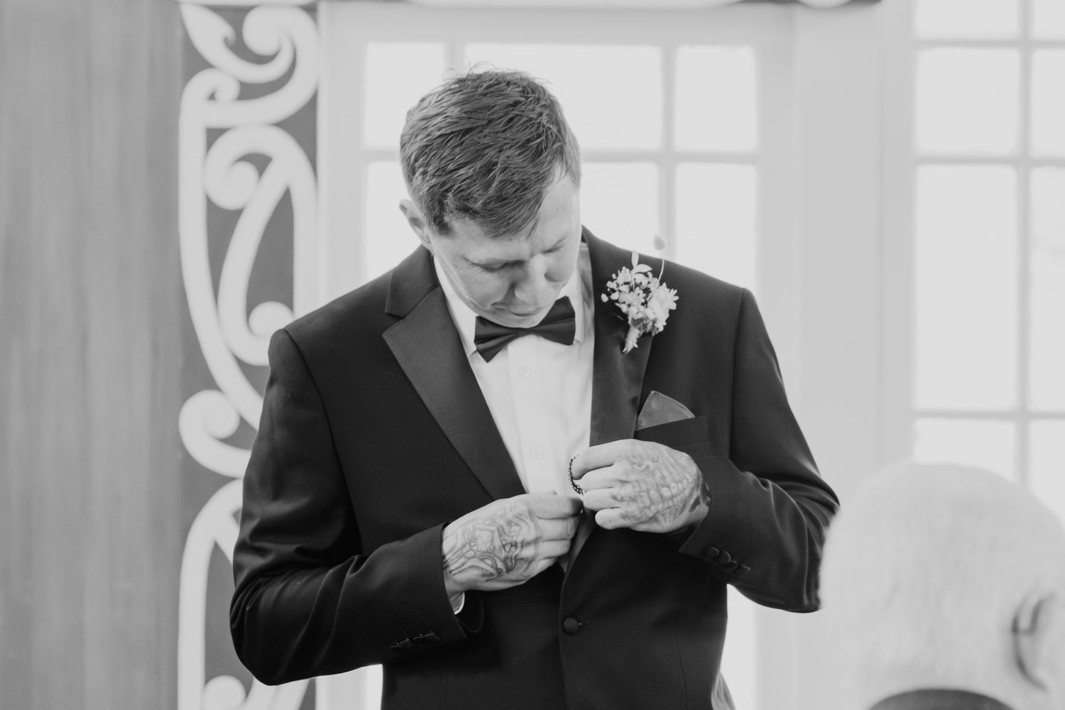 Groom in black tuxedo and bowtie examines something in his hands during formal wedding ceremony.