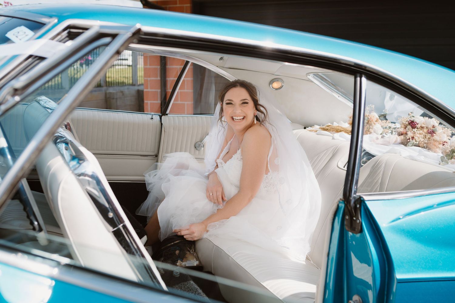 Someone wearing a white wedding dress sits in the driver's seat of a classic turquoise blue vintage car.