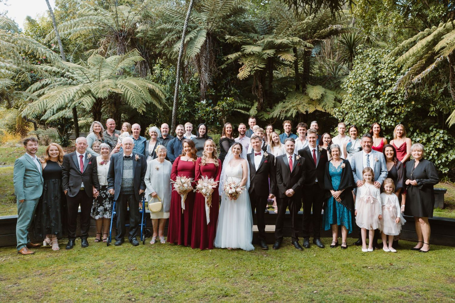 Large wedding party group photo with palm trees and greenery in the background.