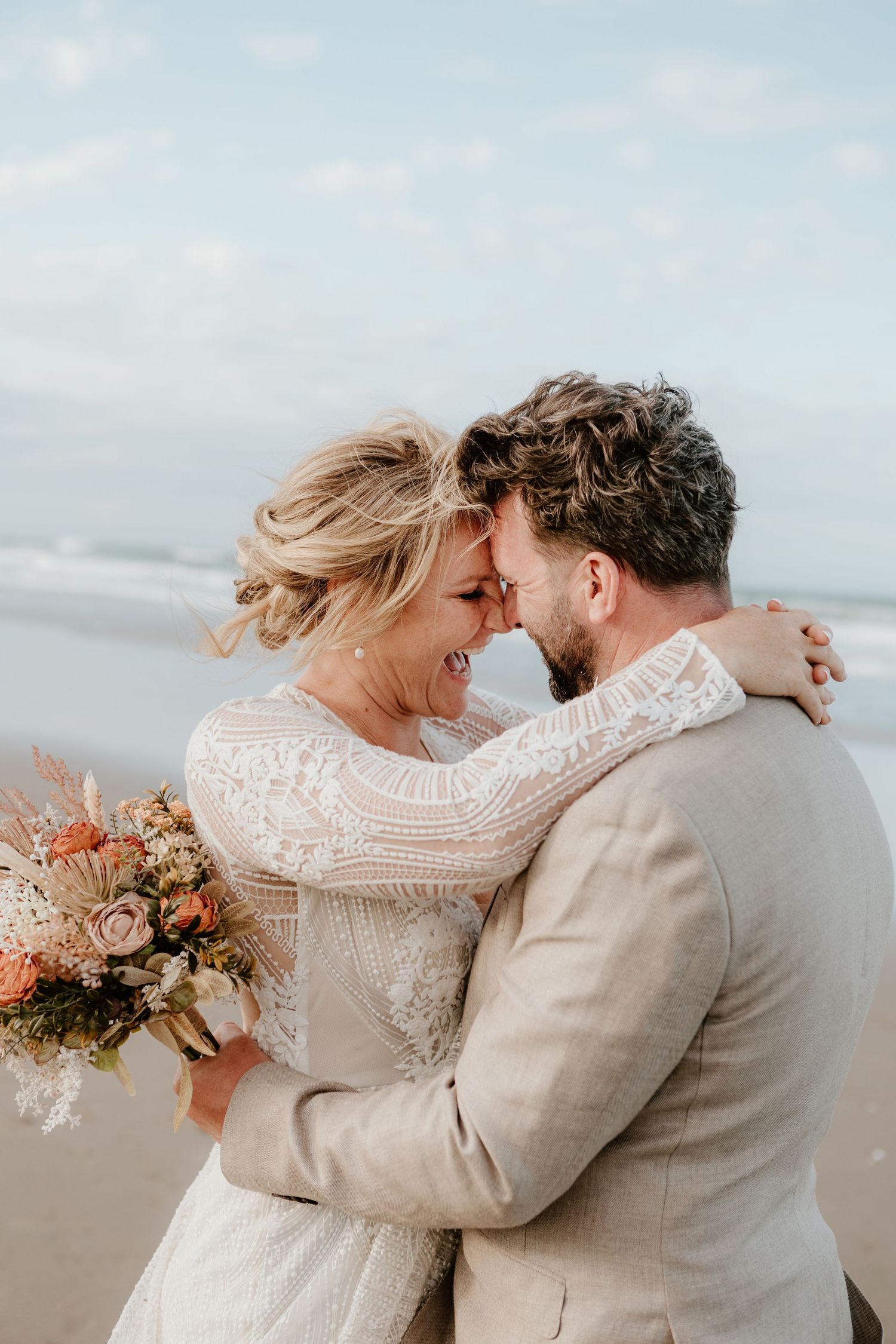 Romantic beach wedding couple embracing with coral and white bouquet on overcast day. Papamoa Surf Lifesaving Club Wedding