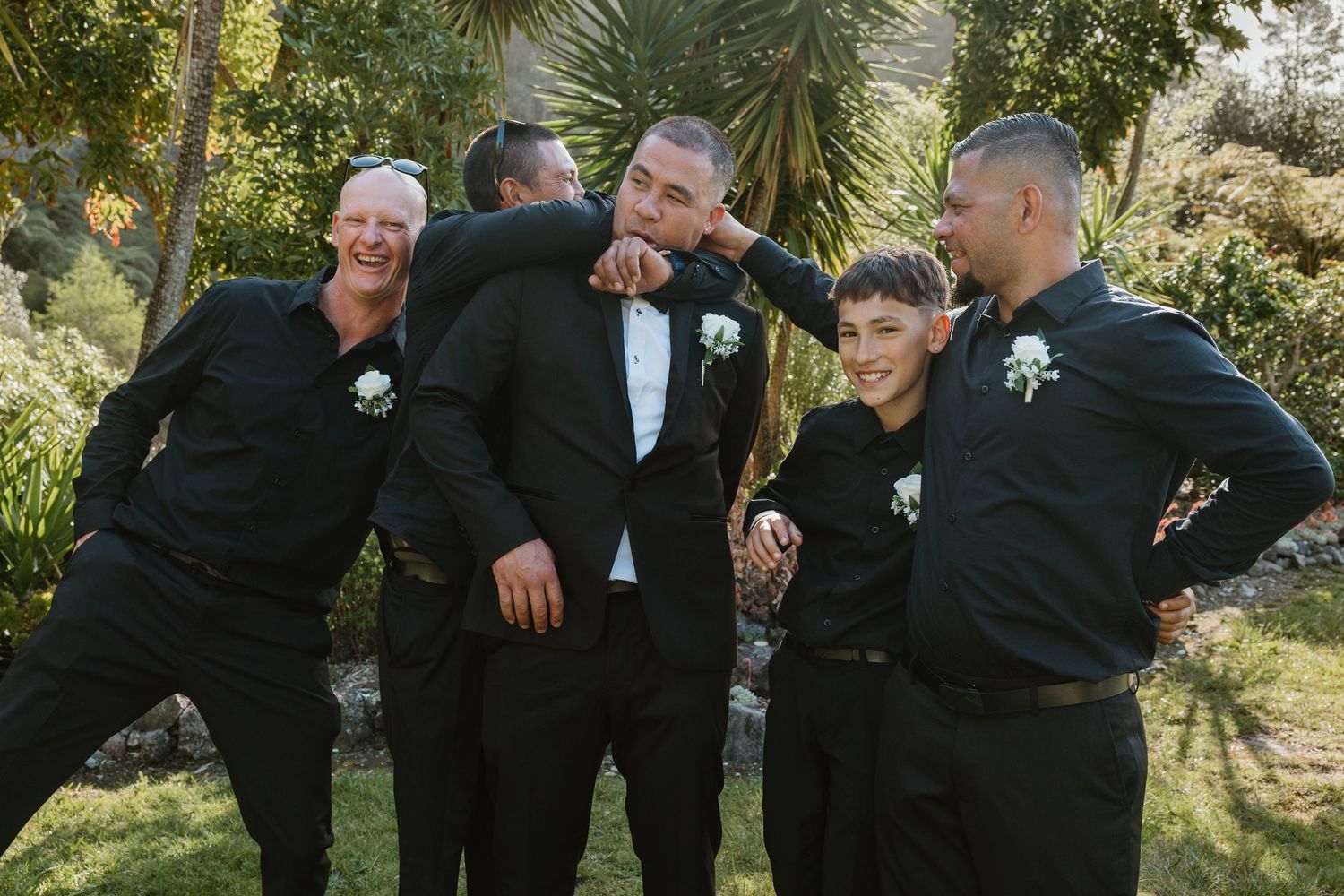 Groomsmen in black suits share a candid moment while laughing and embracing outdoors in a garden setting.