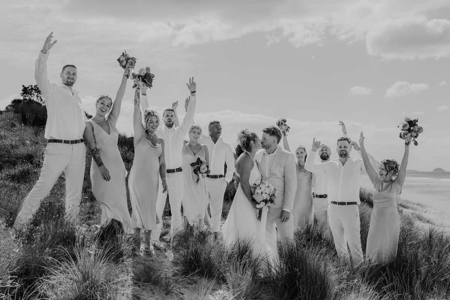 Black and white photo of a joyful wedding party celebrating outdoors near the coast, arms raised in celebration.