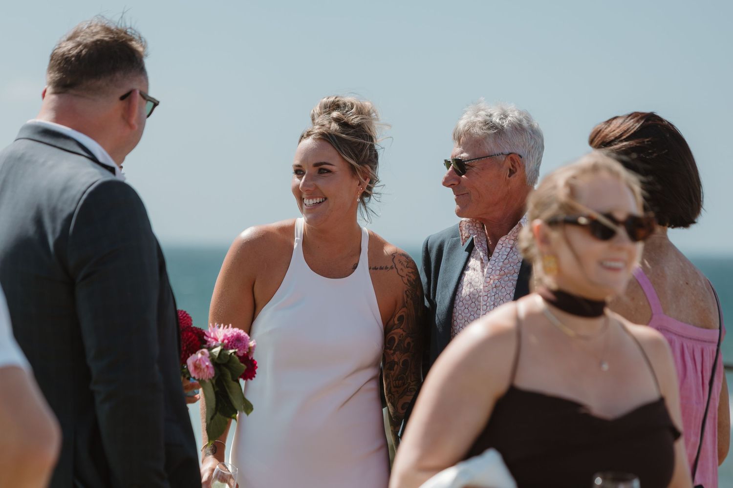 Bride in white dress holding red roses smiles at groom during outdoor beach wedding ceremony with guests.